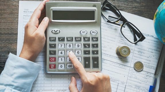 person using calculator to calculate income tax on a desk with paper, glasses and money