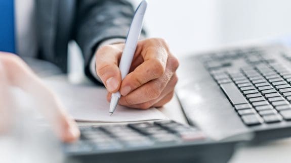 Close up of person holding pen and writing on paper with calculator and keyword in view