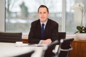 Daniel Gammon is seated at the office table with an open folder, looking ahead.