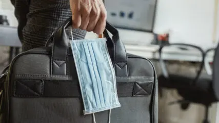 closeup of a young man in an office holding a briefcase and a surgical mask in his hand