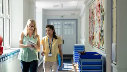 A front view of two female teachers sharing notes and teaching ideas as they walk along the corridor of the school