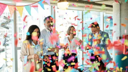 Mature business people celebrating birthday with confetti party popper in office. Group of male and female professionals celebrating birthday party in office.