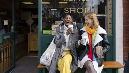 Two girls are sitting outside the cafe, sipping coffee, chatting, and laughing.