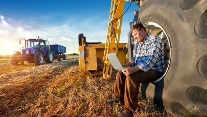 farmer sitting on tractor wheel looking at agricultural technology trends on laptop
