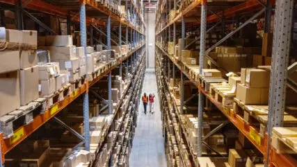 Warehouse shelves filled with boxes, with two workers walking, highlighting supply chain disruption challenges