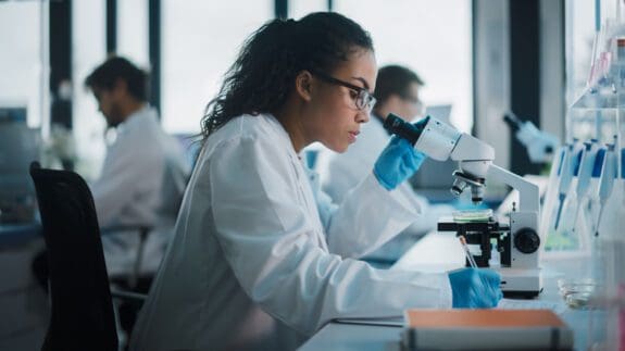 A female scientist in the lab is peering through a microscope.