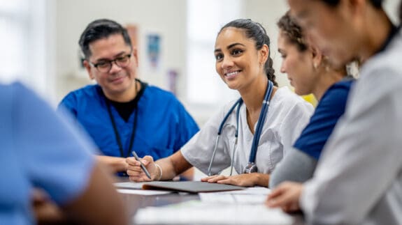 A small medical staff team sit around a boardroom table as they meet to discuss and collaborate on patient cases. They are each dressed professionally and are focused on the meeting.