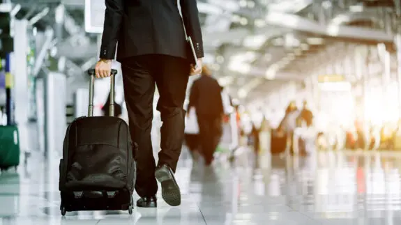 Businessman checking e-mail on mobile phone while walking with suitcase inside airport
