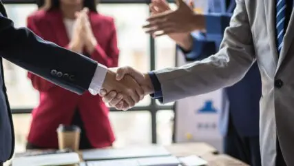 Business men shaking hands in a conference room while people applaud in the background.