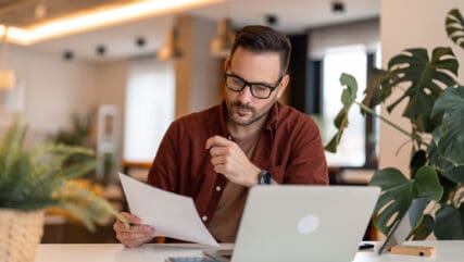 Man sitting at desk looking a VAT inspection papers with laptop on table