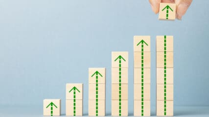 Blue background with blocks stacked up in a line increasing as the go down the line with green arrows pointing to the sky. A hand is placing the final block on the top of the block graph.