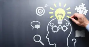 A hand holding chalk using it to draw a lightbulb over a head silhouette on a chalk board.