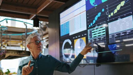 Man in glasses pointing at AI valuations charts and graphs on a digital display