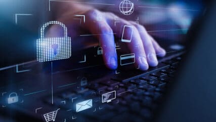 Hand typing on a laptop keyboard with a digital display floating above it. This display includes a digital padlock and other digital icons.