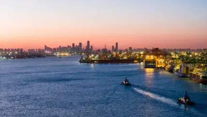 Industrial harbour at sunset with city skyline, glowing port buildings, and boats moving through the water.