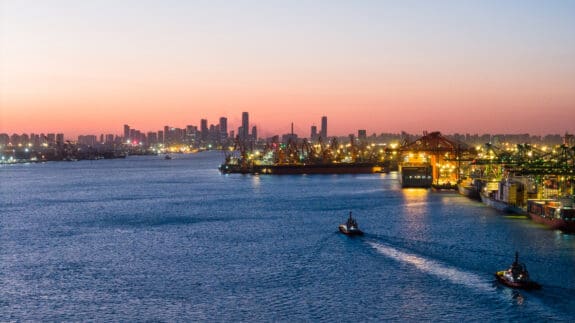 Industrial harbour at sunset with city skyline, glowing port buildings, and boats moving through the water.
