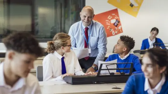 Male teacher with a blue shirt and red tie, teaching a class of secondary school students.