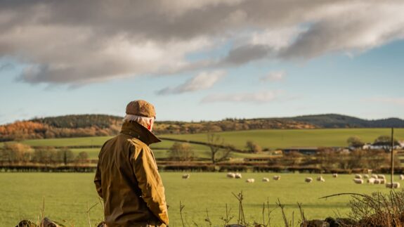 A man with his back to camera wearing a waxed jacket and flat tweed cap looking across a field