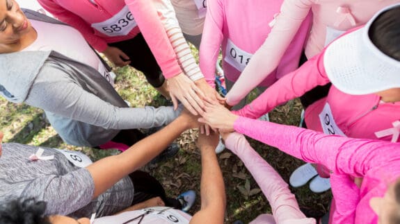 In this view from above, an unrecognizable group of race for the cure participants stand outside in a circle and stack hands.