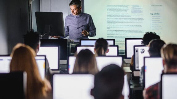 A man stood presenting in front of a group of students with a big screen displayed behind him. The man is presenting on a monitor screen at the front of the room while the class have their monitors open in front of them.