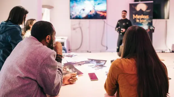 People attending a video game development presentation with concept art on the table and a screen showing gameplay