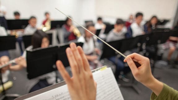 View of band instructor teacher conductor hands leading music performance orchestra of junior high school teenager students practicing in school classroom together in a group of people