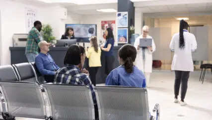 Hospital staff assist patients at front desk in organized emergency waiting area. Nurse in blue scrubs consults seated asian man as other patients and doctors prepare for medical consultations.