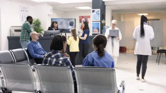 Hospital staff assist patients at front desk in organized emergency waiting area. Nurse in blue scrubs consults seated asian man as other patients and doctors prepare for medical consultations.