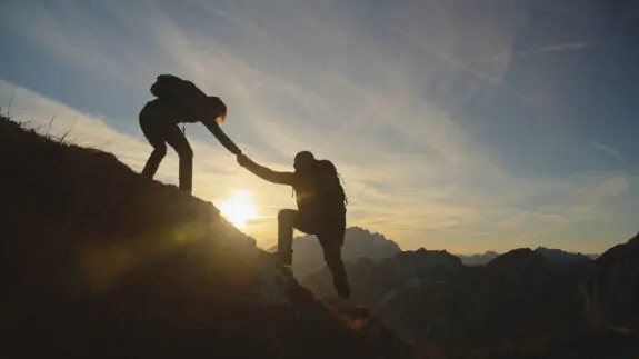 A mid-adult man and woman clad in hiking attire exchange a joyful high five on a mountaintop, silhouetted against a serene sunset sky.