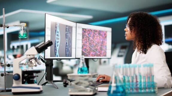 Female medical doctor in a lab using a computer for DNA research. Test tubes and a microscope can be seen in the foreground.