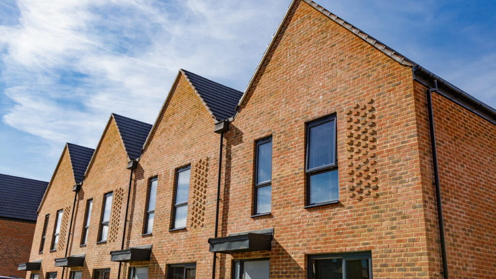 Colour image depicting a brand new housing estate and construction - new houses are still being built on the site. The rows of houses include terraced housing and detached, with pristine green lawns and gardens.