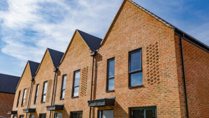 Colour image depicting a brand new housing estate and construction - new houses are still being built on the site. The rows of houses include terraced housing and detached, with pristine green lawns and gardens.
