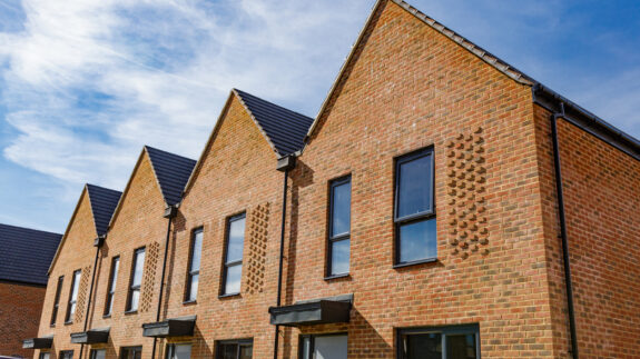 Colour image depicting a brand new housing estate and construction - new houses are still being built on the site. The rows of houses include terraced housing and detached, with pristine green lawns and gardens.