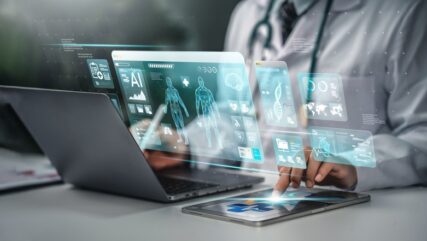 A doctor using a tablet and a laptop, with a holographic overlay showing a human body, DNA, and reports.